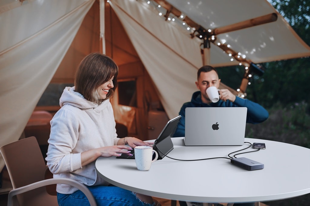 Un homme et une femme travaillant sur un ordinateur portable et une tablette, connectés à un hub, assis devant une tente de glamping éclairée