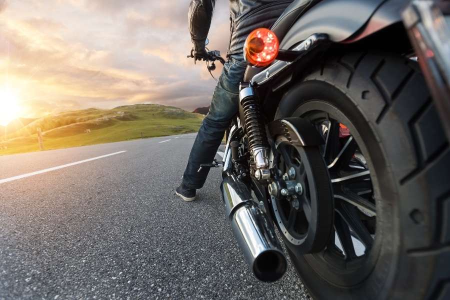 Vue arrière d'un motard en jean et veste de cuir sur une moto à forte cylindrée de moto, avec échappement chromé et large pneu arrière, sur une route au coucher du soleil.