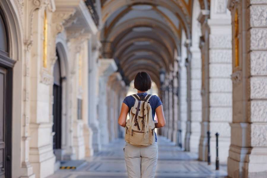 Une voyageuse avec un sac à dos beige marche seule sous les arcades d'un bâtiment historique, illustrant les erreurs de voyage liées au manque de planification.
