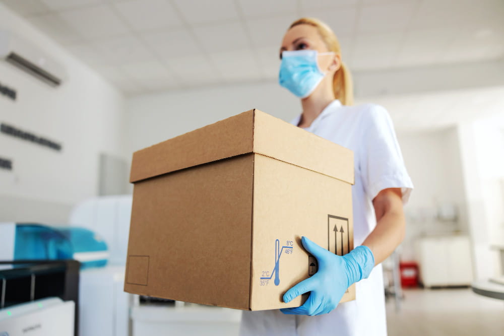 Une professionnelle de santé portant un masque et des gants transporte un carton sécurisé lors d'un déménagement de laboratoire