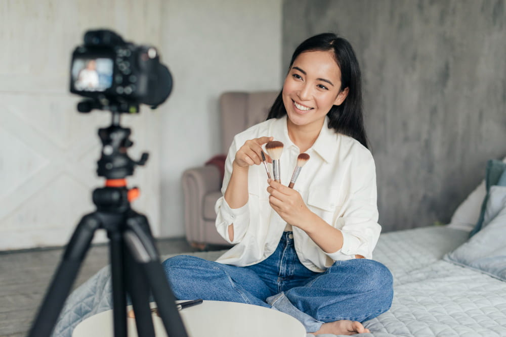 Une jeune femme souriante assise sur son lit présente des pinceaux de maquillage devant sa caméra sur trépied lors d'une session de vlogging dans sa chambre.