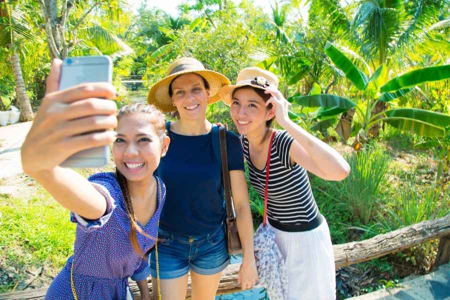 Trois jeunes femmes, dont deux portent des chapeaux de paille, sourient largement en prenant un selfie avec un smartphone dans un jardin tropical luxuriant, un lieu typique des pièges à touristes.